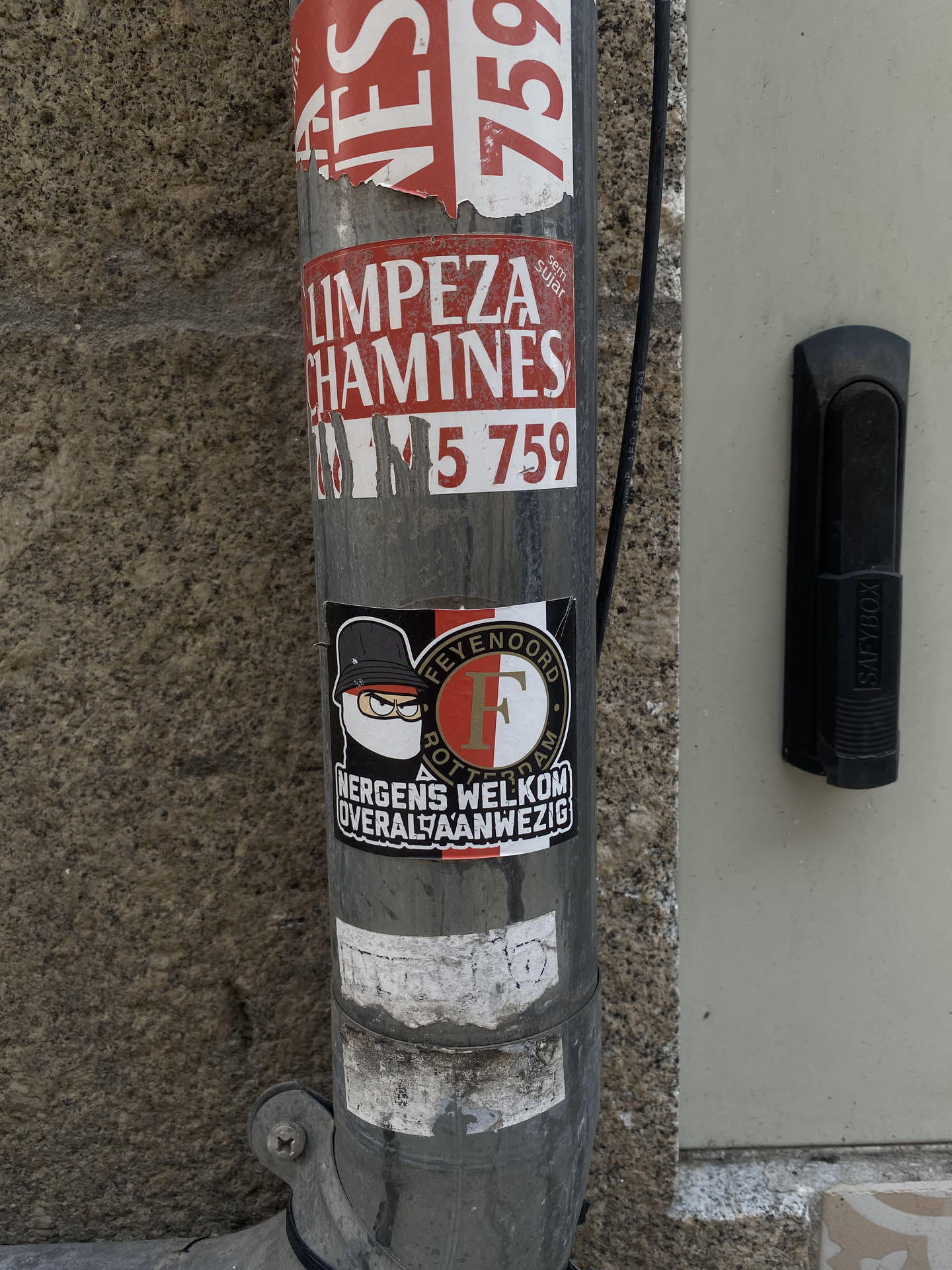 A rectangular sticker featuring a stylized cartoon character on the left and a circular football club logo on the right, set against a predominantly black background. The character wears a black bucket hat, yellow-rimmed glasses, and a white face mask, with a stern expression. The circular logo is split vertically into red and white halves, with a large white 'F' in the center. The words 'FEYENOORD' and 'ROTTERDAM' are written in gold/yellow text around the 'F' within a black outer ring. Below the character, the text 'NERGENS WELKOM' is in white, and below the logo, 'OVERAL AANWEZIG' is also in white, both on a black background.