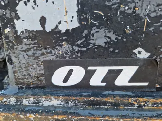 A close-up shot of a black sticker with the letters "OTL" in white. The sticker is affixed to a heavily textured, weathered surface, possibly metal, with peeling paint and various markings.
