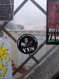 Street sticker The sticker is circular with a black background. It features a stylized illustration of a person wearing a dark hoodie and a balaclava, holding a megaphone in one hand and a red object (possibly a flare or baton) in the other. The text 'YNGRE ULTRAS SANDEFJORD' is written in white capital letters around the top edge of the circle, and 'YUS' is written in large white capital letters at the bottom center.