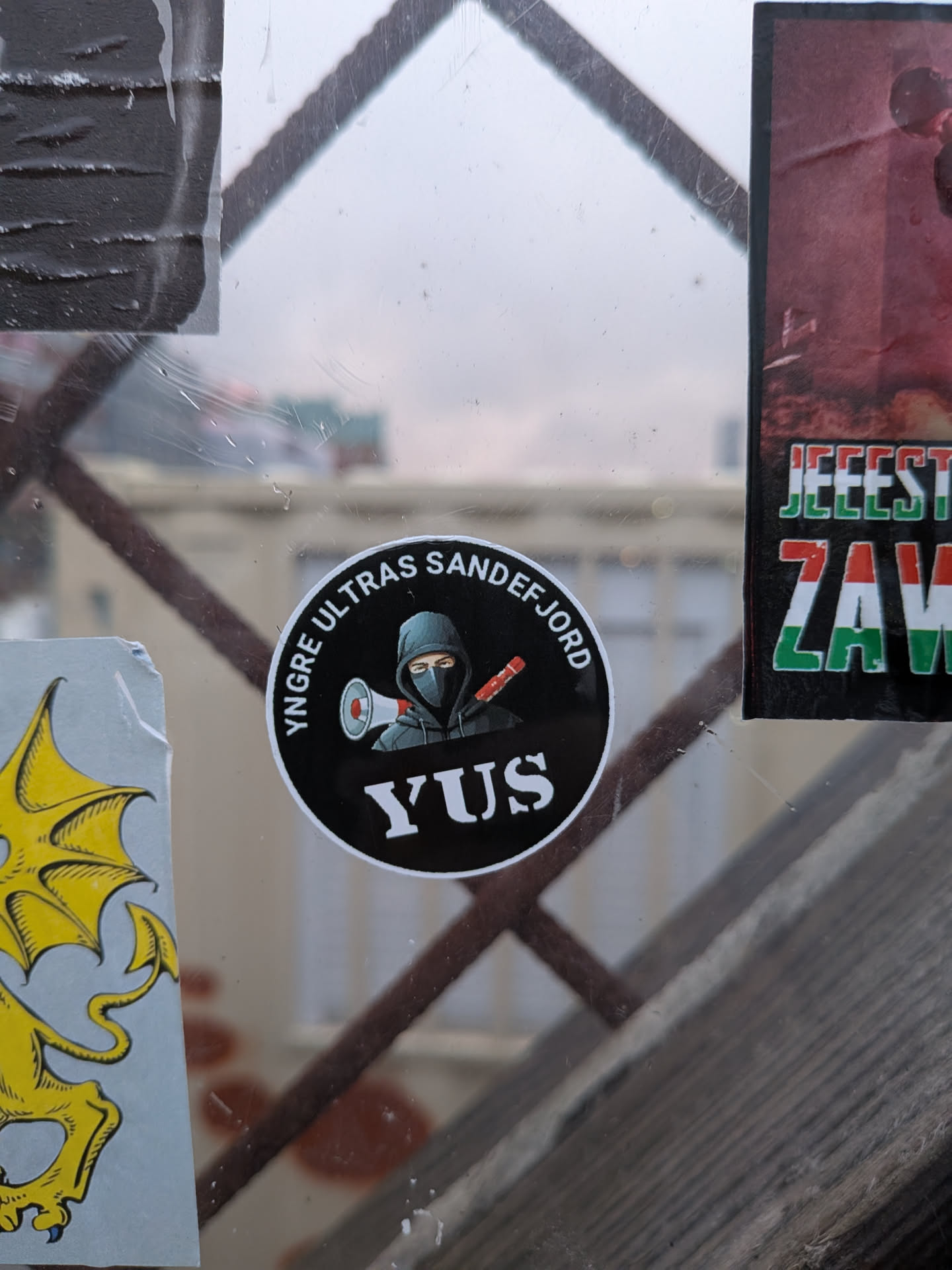 Street sticker The sticker is circular with a black background. It features a stylized illustration of a person wearing a dark hoodie and a balaclava, holding a megaphone in one hand and a red object (possibly a flare or baton) in the other. The text 'YNGRE ULTRAS SANDEFJORD' is written in white capital letters around the top edge of the circle, and 'YUS' is written in large white capital letters at the bottom center.