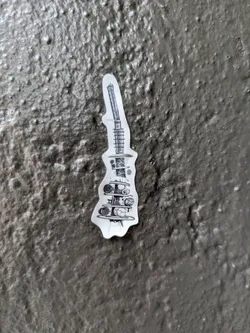 Street sticker A vertically elongated sticker featuring a detailed black and white line drawing. The artwork depicts a complex, almost surreal structure that resembles a cross-section of a futuristic machine or a deconstructed architectural element. It includes a tower-like top section with linear details, possibly representing a spring or antenna, leading down to multiple platforms. These platforms hold various spherical or orb-like objects, some appearing cracked or broken, suggesting decay or fragmentation. The overall aesthetic is one of intricate, almost blueprint-like design with a distressed or post-apocalyptic feel. The sticker is placed on a rough, dark grey or black textured surface.