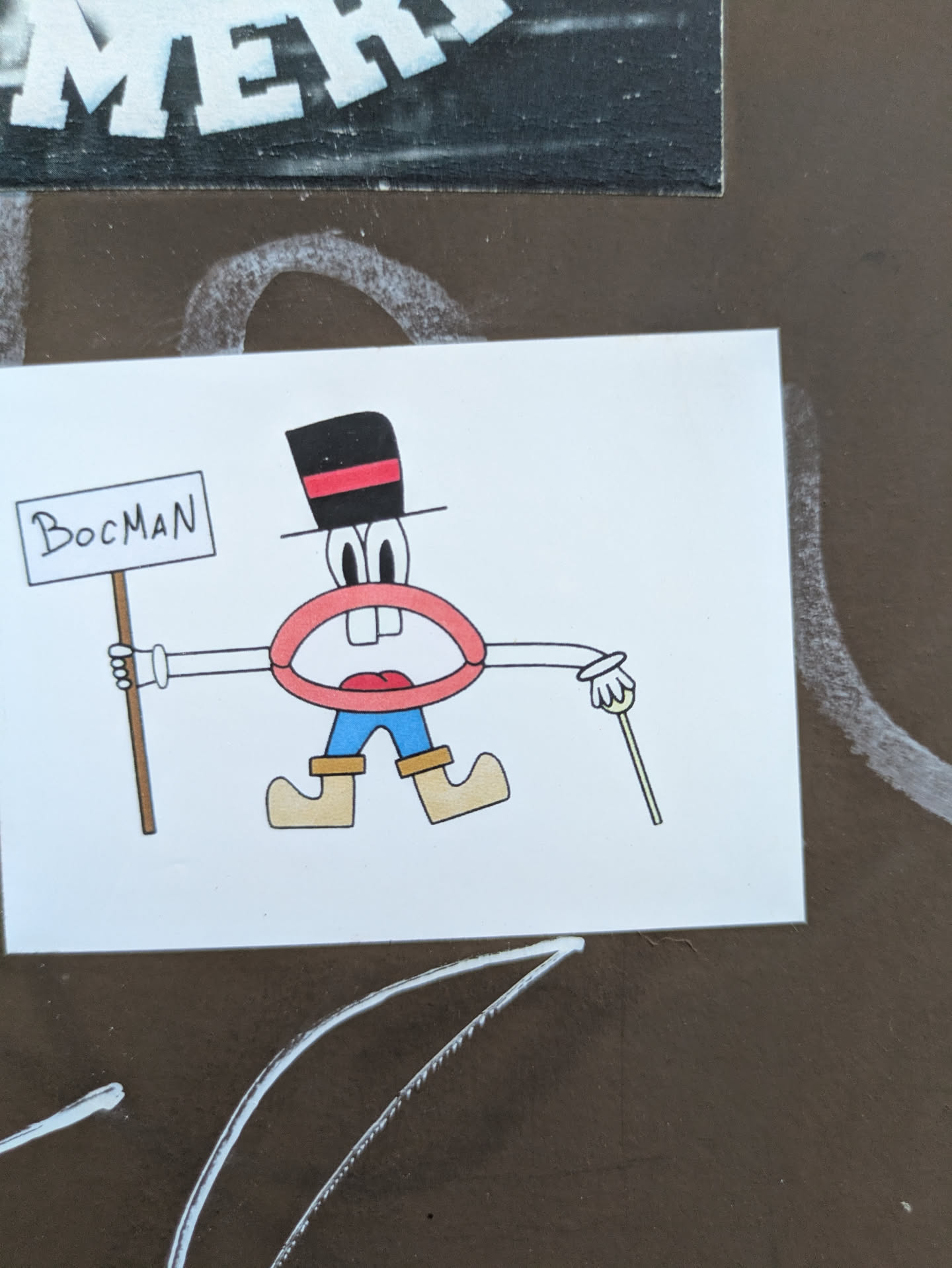 A cartoonish character sticker on a wall. It's a mouth with eyes and a black top hat with a red stripe, wearing blue trousers and brown shoes, holding a sign that says "BocMan" in one hand and a cane in the other.
