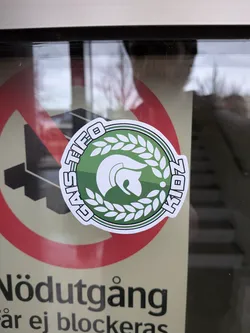 Street sticker The sticker is circular with a white border and a black outline. The central design features a green gradient background. Within this, a white stylized helmet, reminiscent of a Spartan or Roman helmet, is depicted. The helmet is surrounded by a white laurel wreath. Along the top edge of the circle, in white block letters with a black outline, is the text 'GAS TIFO'. Along the right edge, also in white block letters with a black outline, is the year '2014'. The sticker has a slightly glossy finish and appears to be adhered to a glass surface, as evidenced by reflections.