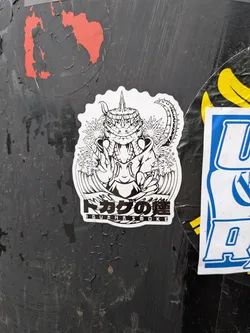 The sticker is a black and white illustration with an irregular, wavy white border. The central figure is a highly stylized, anthropomorphic iguana or lizard creature with sharp features, including prominent teeth, intense eyes, and a spiky crown or headpiece. Its body is depicted wearing what appears to be a hooded garment, and a spiky tail is visible on the right. Flanking the creature are detailed cannabis leaves, and below it, large, dynamic waves are rendered in a style reminiscent of traditional Japanese Ukiyo-e art. At the bottom, a black rectangular bar contains white text, both in Japanese characters and English.