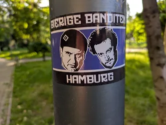 Street sticker This is a sticker featuring black and white portraits of two men against a blue background, with the words "BIERIGE BANDITEN" above and "HAMBURG" below. The sticker is affixed to a gray metal pole in an outdoor setting.