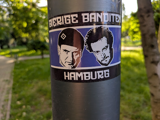 This is a sticker featuring black and white portraits of two men against a blue background, with the words "BIERIGE BANDITEN" above and "HAMBURG" below. The sticker is affixed to a gray metal pole in an outdoor setting.