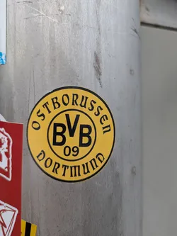 Street sticker The sticker is circular, featuring a vibrant yellow background with a thick black outer border. In the center, there is a smaller black circle containing the bold black letters "BVB" with "09" directly underneath. Around this central emblem, along the outer edge of the yellow circle, the word "OSTBORUSSEN" is written in black uppercase letters across the top, and "DORTMUND" is written in similar black uppercase letters across the bottom. The overall design is clean and clearly identifies with the football club.
