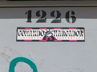 Street sticker A rectangular sticker with the text "Comando Marcador Levante U.E. Supporters" is affixed to a grey surface, with the number "1226" stenciled above it. The sticker features a cartoon mascot. There is some green paint below the sticker.