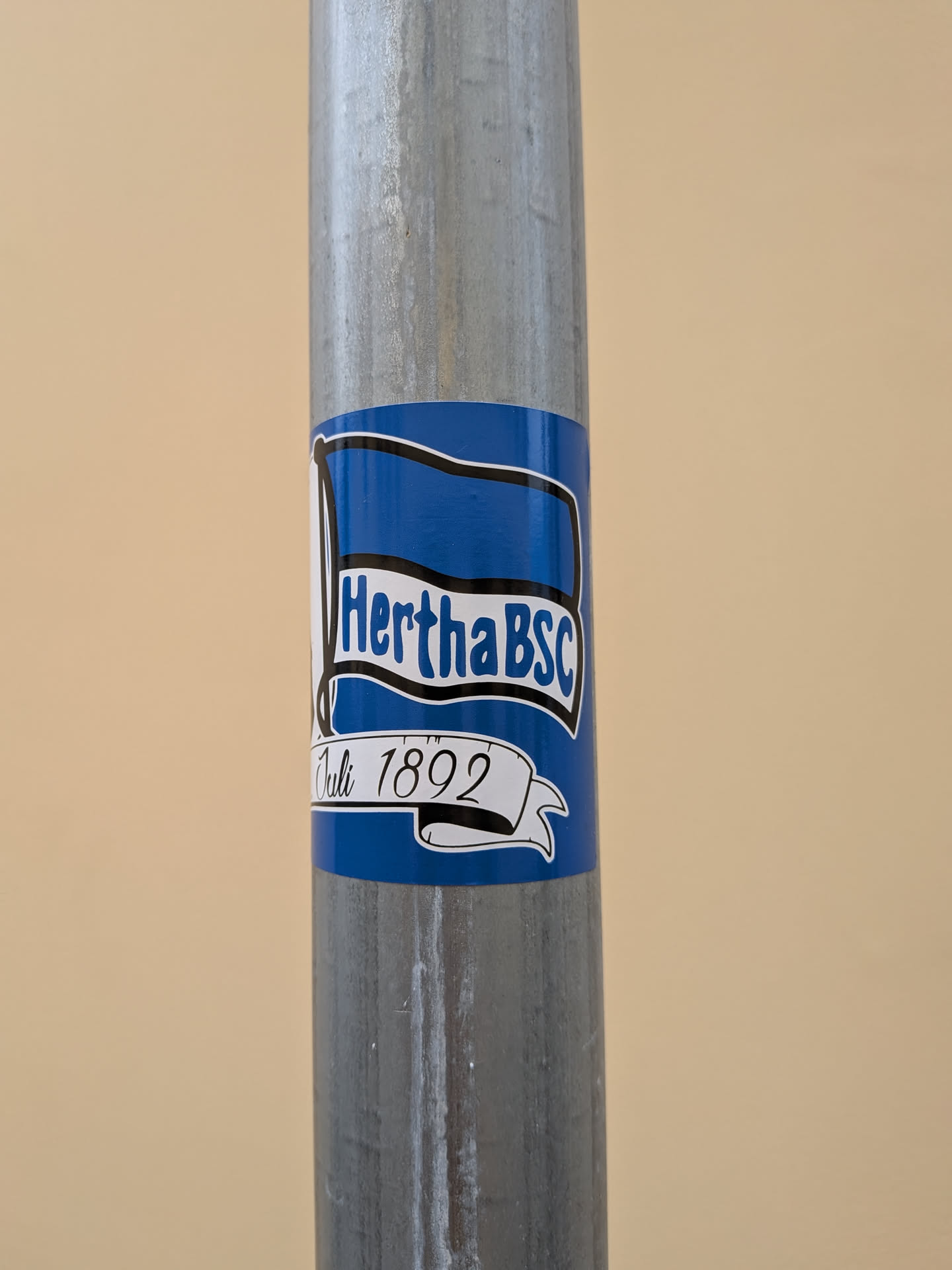 A blue sticker featuring the Hertha BSC blue-and-white flag logo. Below the flag, a white banner reads 'Juli 1892' in black script. The sticker is wrapped around a galvanized metal pole.