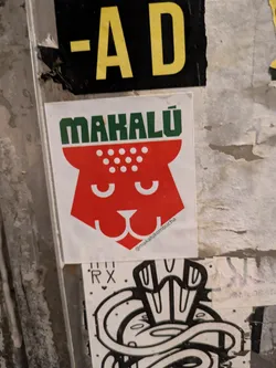 Street sticker This sticker promotes Mahalú kombucha. It features a simplified, red jaguar head design with white dots on top. The brand name 'MAHALÚ' is written in green above the head. The text '@makalukombucha' is on the bottom right of the sticker.