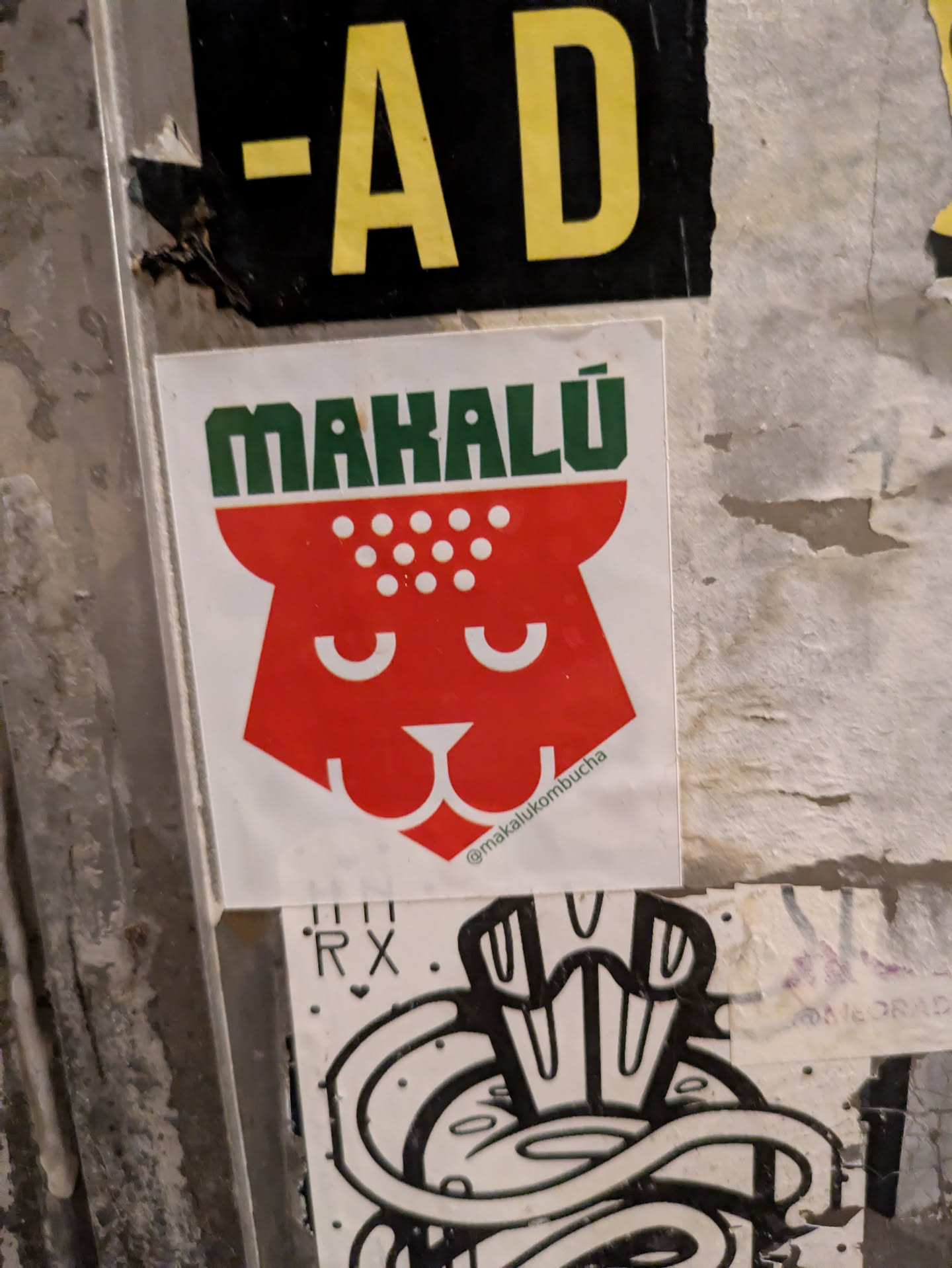 This sticker promotes Mahalú kombucha. It features a simplified, red jaguar head design with white dots on top. The brand name 'MAHALÚ' is written in green above the head. The text '@makalukombucha' is on the bottom right of the sticker.