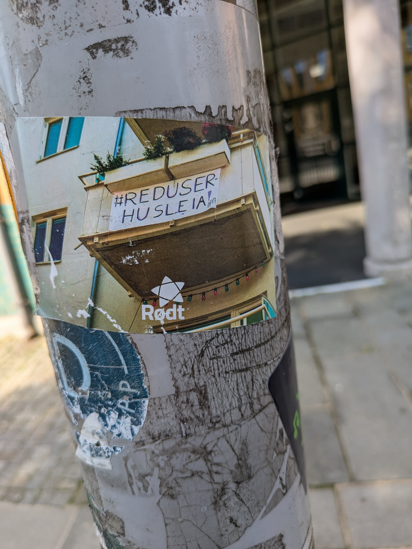 The sticker depicts a photograph of a building's balcony, from which a white banner is hung, displaying the text '#REDUSER-HUSLEIA!'. Below the balcony, a string of colorful lights is visible. At the bottom of the sticker, the word 'Rødt' is prominently displayed next to a stylized white star, which is the logo associated with the Norwegian political party, Rødt. The sticker itself is affixed to a heavily worn and layered pole in an urban setting.