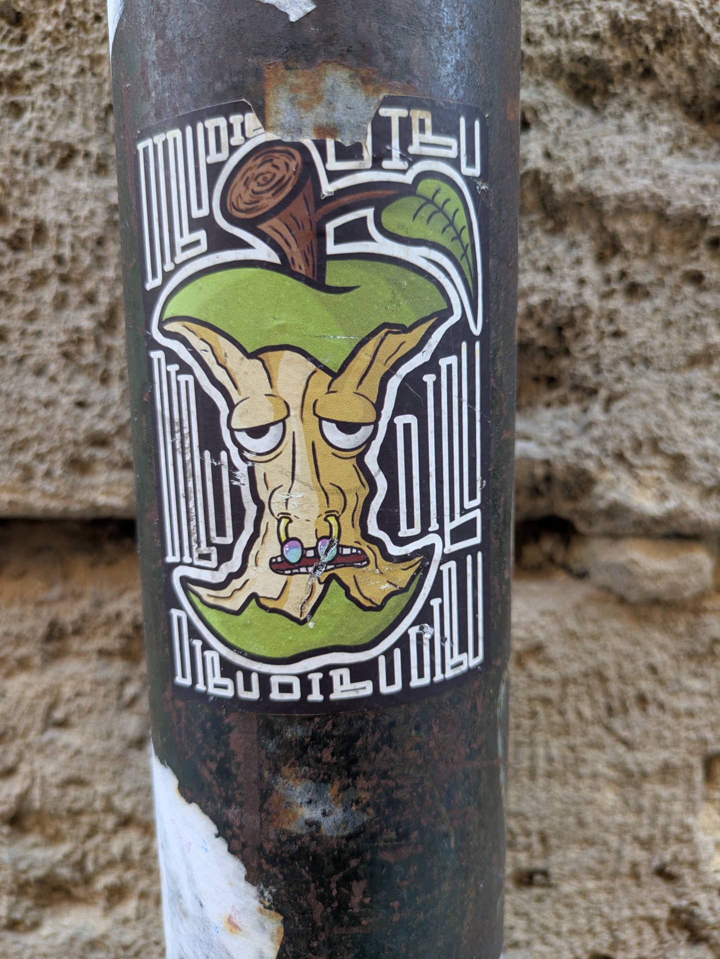A unique sticker featuring an apple core with a whimsical, somewhat grotesque face, placed against a decorative backdrop of vertical lines. The apple core has a grumpy expression and unusual facial features, including multi-colored nostrils and a generally weathered look. The sticker is adhered to a rusty metal pole.