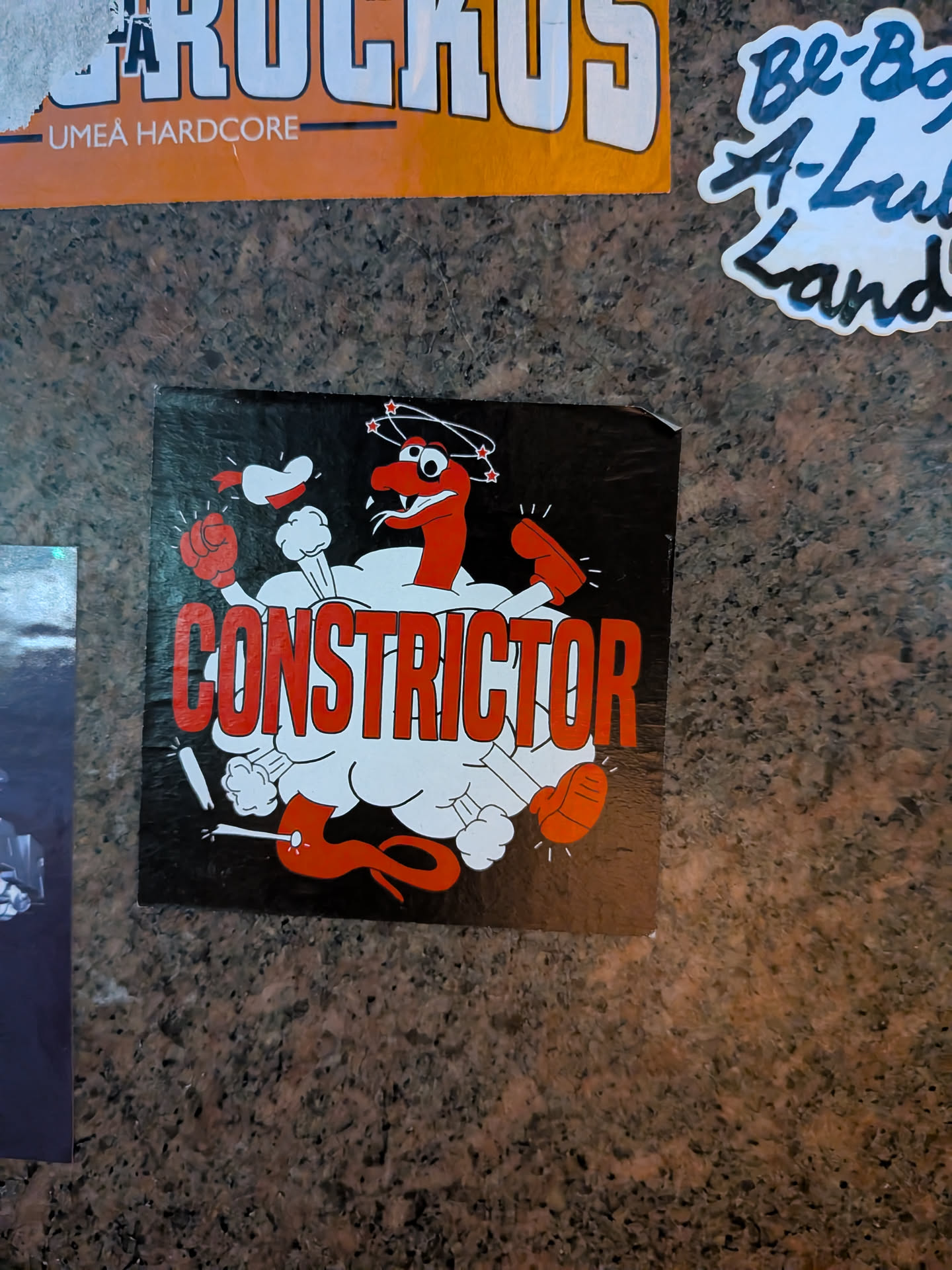 The sticker features a black background with a cartoonish red snake character. The snake has a long, segmented body and a head with wide eyes and a slightly open mouth, with three red stars circling its head, indicating dizziness. It is depicted in a dynamic pose, seemingly emerging from or surrounded by white, puffy clouds or smoke. The snake is wearing red boxing gloves on its front limbs and has red feet. The word 'CONSTRICTOR' is prominently displayed in large, bold, red, block letters across the center of the sticker, partially obscured by the white cloud-like elements. The overall style is reminiscent of vintage cartoons or comic books.