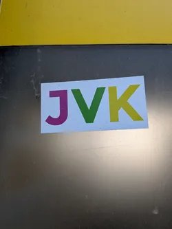 Street sticker The sticker is a rectangular shape with a clean white background. It prominently displays three uppercase letters: 'J', 'V', and 'K'. The 'J' is a vibrant magenta or purple, the 'V' is a bright green, and the 'K' is a golden yellow. The letters are sans-serif, bold, and evenly spaced, filling most of the sticker's width. The sticker appears to be applied to a dark, possibly metallic, surface and shows minor signs of wear, such as small specks and slight texture on its surface.