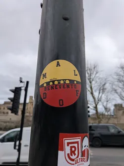 Street sticker A circular sticker adhered to a black pole, distinctly divided into two horizontal color sections: a bright yellow top half and a deep red bottom half. A black silhouette of an arched bridge spans the horizontal divide, with several small, stylized figures standing atop the bridge, appearing to celebrate or hold hands. The word 'BENEVENTO' is prominently displayed in bold black capital letters across the middle of the sticker, partially overlapping the bridge. Around the upper curve of the yellow section, the letters 'M A L' are spaced out in black. Similarly, around the lower curve of the red section, the letters 'D O F' are spaced out in black. The sticker exhibits minor wear, consistent with outdoor placement.