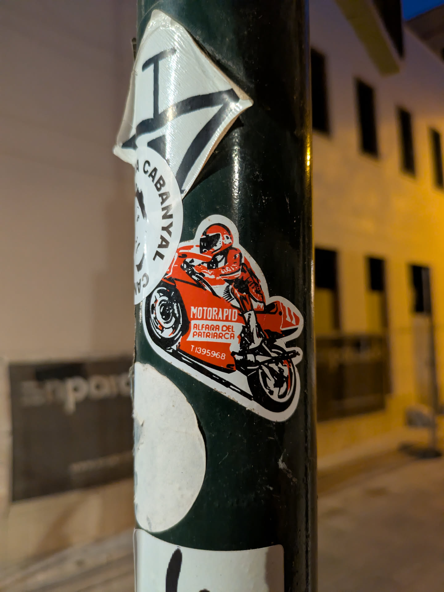 Street sticker This is a photo of a Motorapid sticker that is placed on a dark green pole. The sticker shows a red motorcycle rider. The text on the sticker reads 'MOTORAPID Alfara del Patriarca T.1395968'
