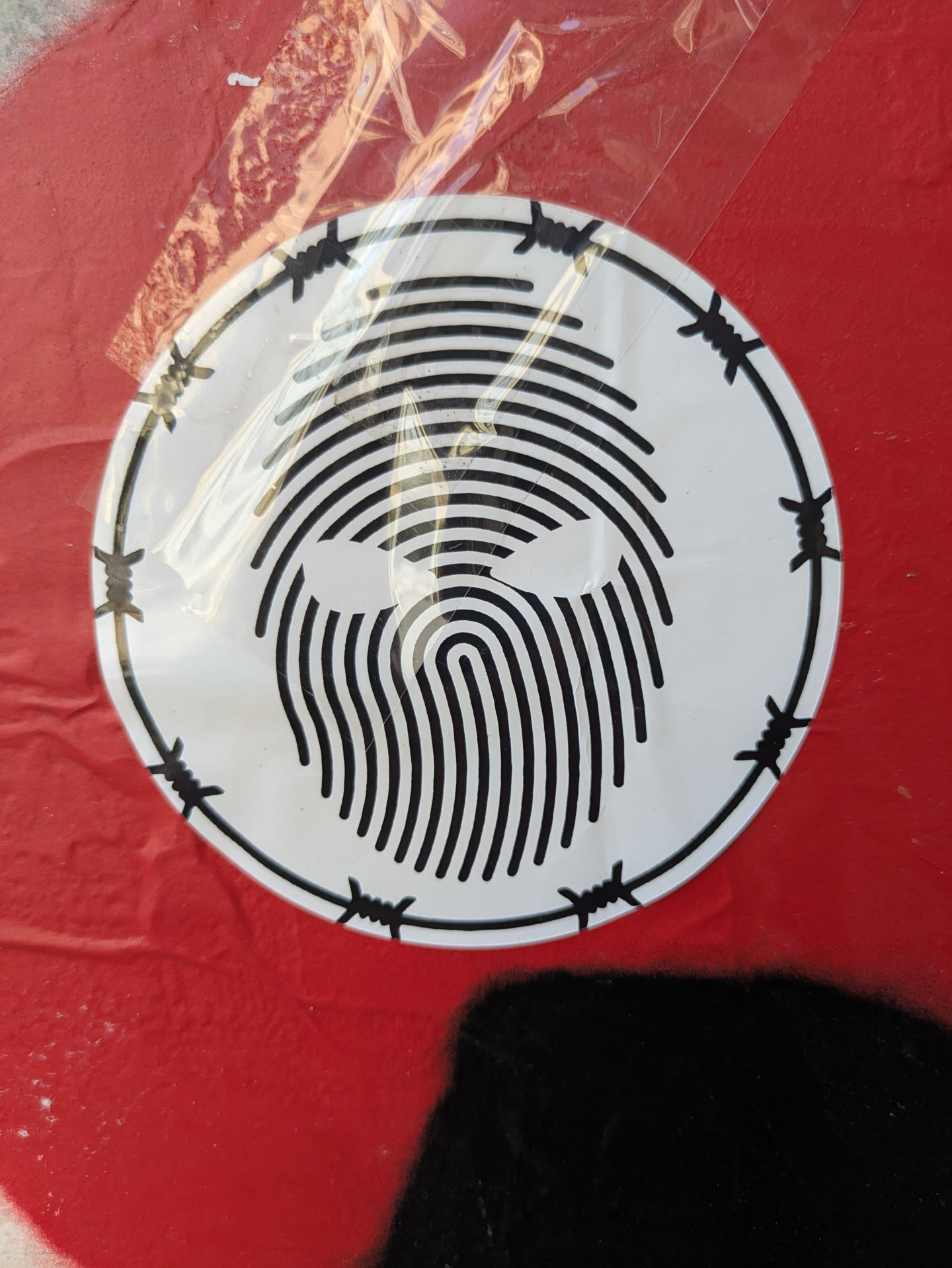 A white circular sticker is adhered to a red surface. The sticker features a design that resembles a fingerprint pattern in the center, made up of black lines. The design is encircled by a black border decorated with barbed wire illustrations. The sticker has some wrinkles.