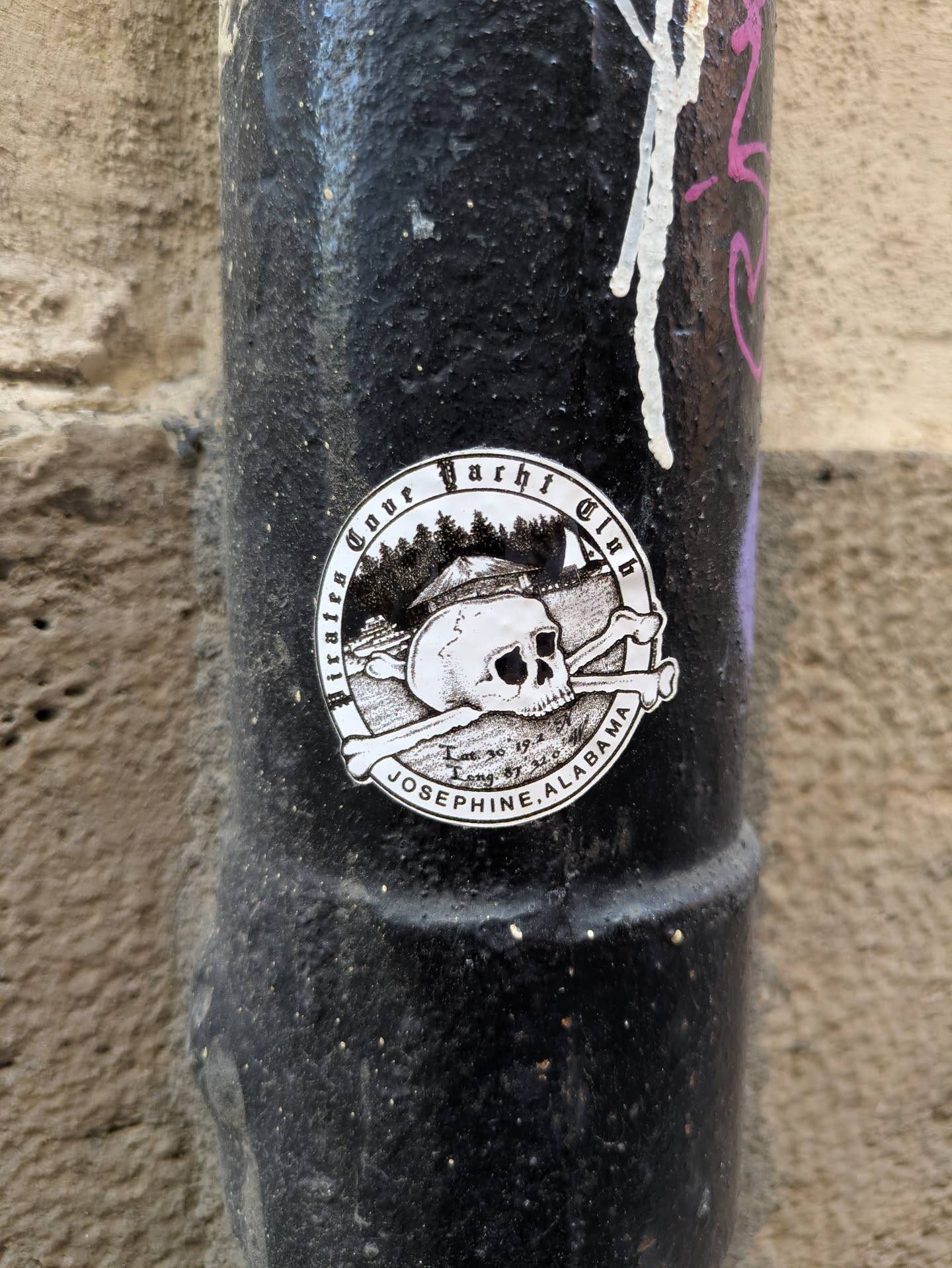 A round sticker affixed to a black pole features a stylized skull and crossbones emblem within a circular border. Above the skull, there is a landscape with trees and what appears to be a small building. The sticker reads "Pirates Cove Yacht Club" along the top border and "Josephine, Alabama" along the bottom. Coordinates "Lat 30° 19.4" and "Long 87° 32.6" are faintly visible beneath the skull.