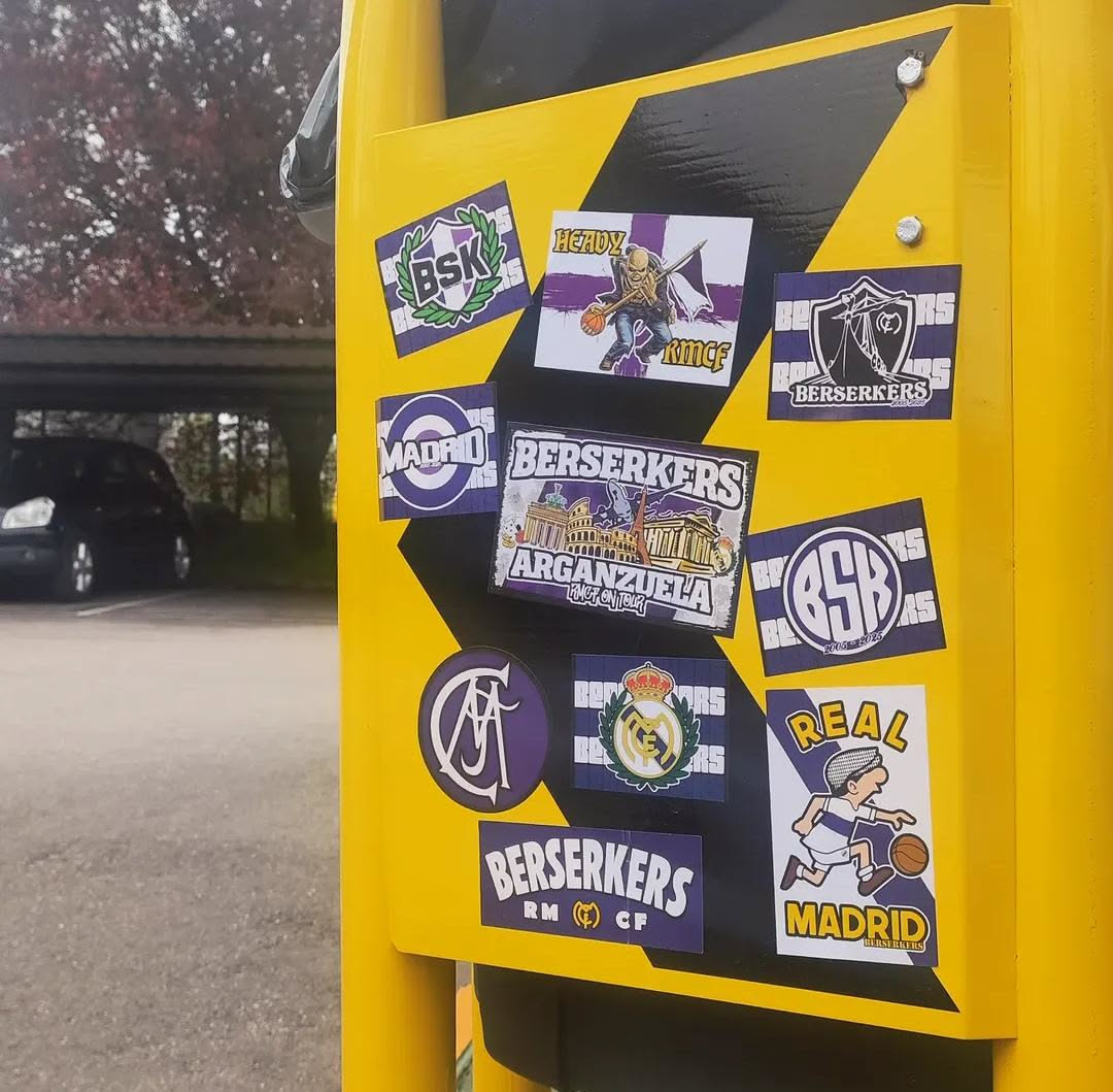 A cluster of various stickers, predominantly in shades of purple, white, and green, affixed to a yellow surface with black diagonal stripes. The stickers feature a mix of club crests, text, and cartoon illustrations, all centered around sports and fan culture, particularly basketball and Madrid-related themes. Key elements include multiple 'BERSERKERS' logos, 'MADRID' text, a figure playing basketball, a crest resembling the Real Madrid logo, and architectural landmarks like the Colosseum. The overall aesthetic is that of a passionate sports fan's collection.