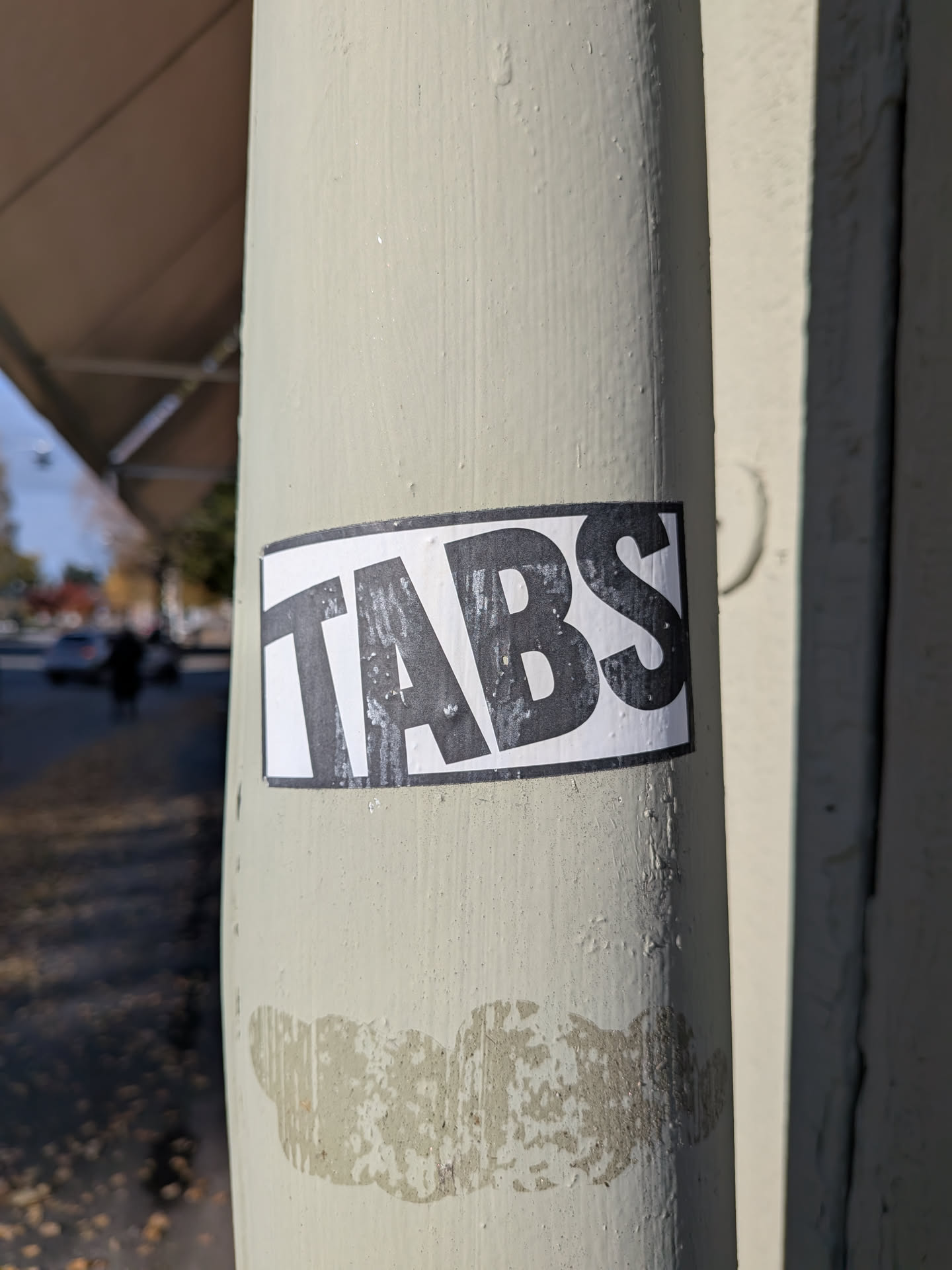 The sticker is rectangular with a white background and a thick black border. The word 'TABS' is printed in large, bold, black sans-serif capital letters. The sticker appears slightly worn, with some scuff marks and imperfections on its surface and edges, suggesting it has been exposed to the elements.