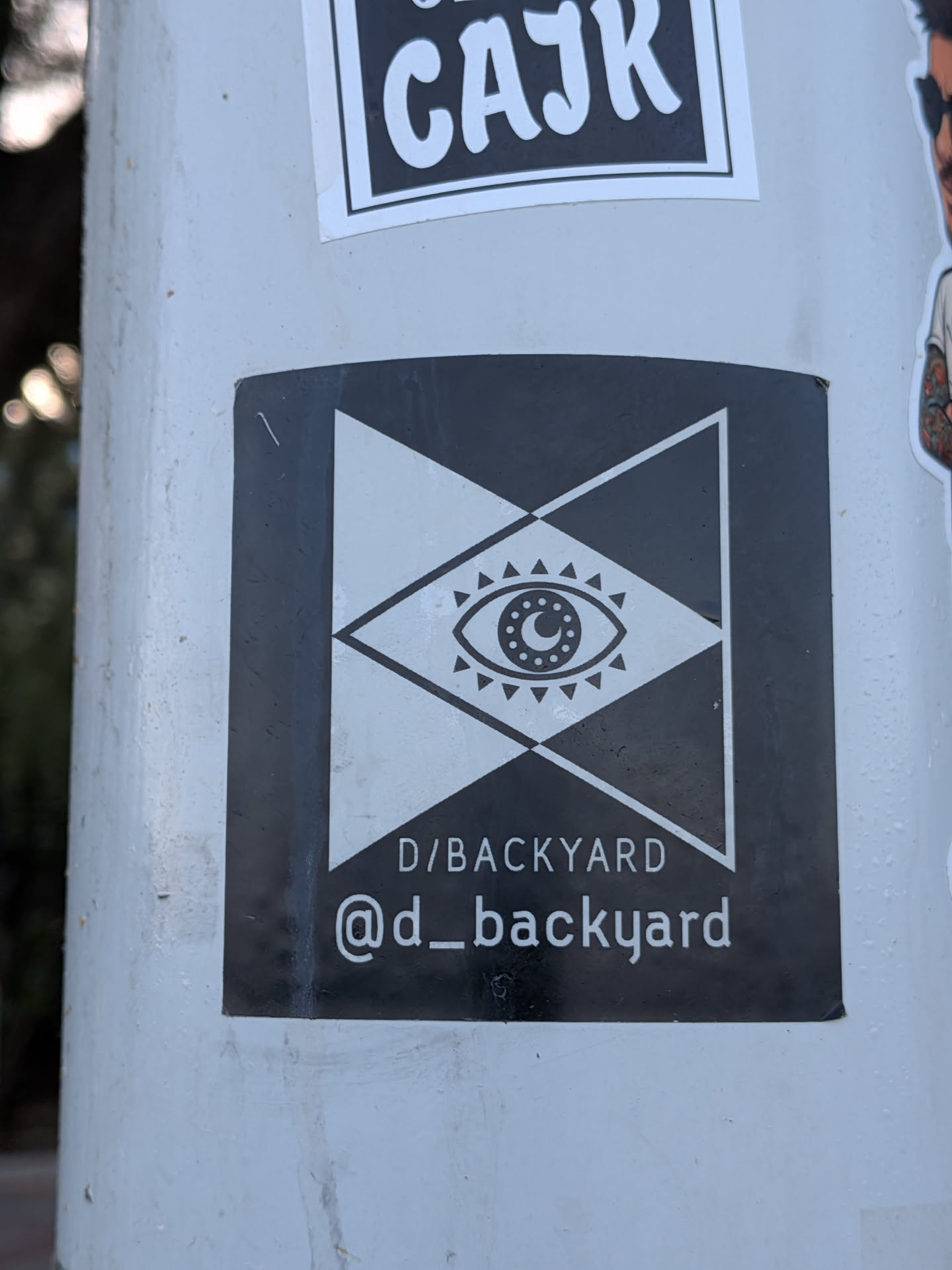 A black rectangular sticker featuring a white geometric design of intersecting triangles framing a central eye with a crescent moon pupil. Text at the bottom reads 'D/BACKYARD' and '@d_backyard'.