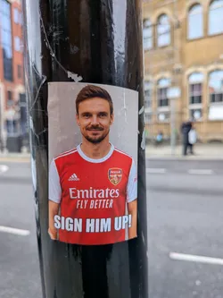 Street sticker A rectangular, vertically oriented sticker featuring a smiling man with short brown hair and a light beard, wearing a red and white Arsenal home jersey. The Adidas logo is visible on the right chest, and the Arsenal crest is on the left. Uniquely, the traditional cannon within the Arsenal crest has been replaced with a small, circular photo of a blonde woman. Below the crest, the main sponsor 'Emirates FLY BETTER' is displayed in white text. At the bottom of the sticker, in large white capital letters on a red background, is the phrase 'SIGN HIM UP!'. The background behind the man is a plain, light grey or beige color, suggesting a wall. The sticker is adhered to a black pole, showing some minor creases and wear around its edges.
