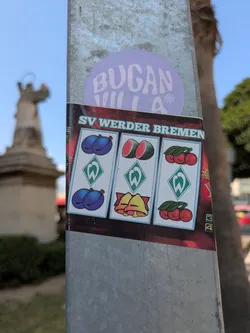 Street sticker A rectangular sticker attached to a pole featuring a slot machine design with "SV Werder Bremen" at the top. The slot reels display images of plums, watermelons, cherries, bells and the SV Werder Bremen logo. The sticker is colorful and eye-catching. Above it is a round purple sticker with the word "BUGANVILLA".