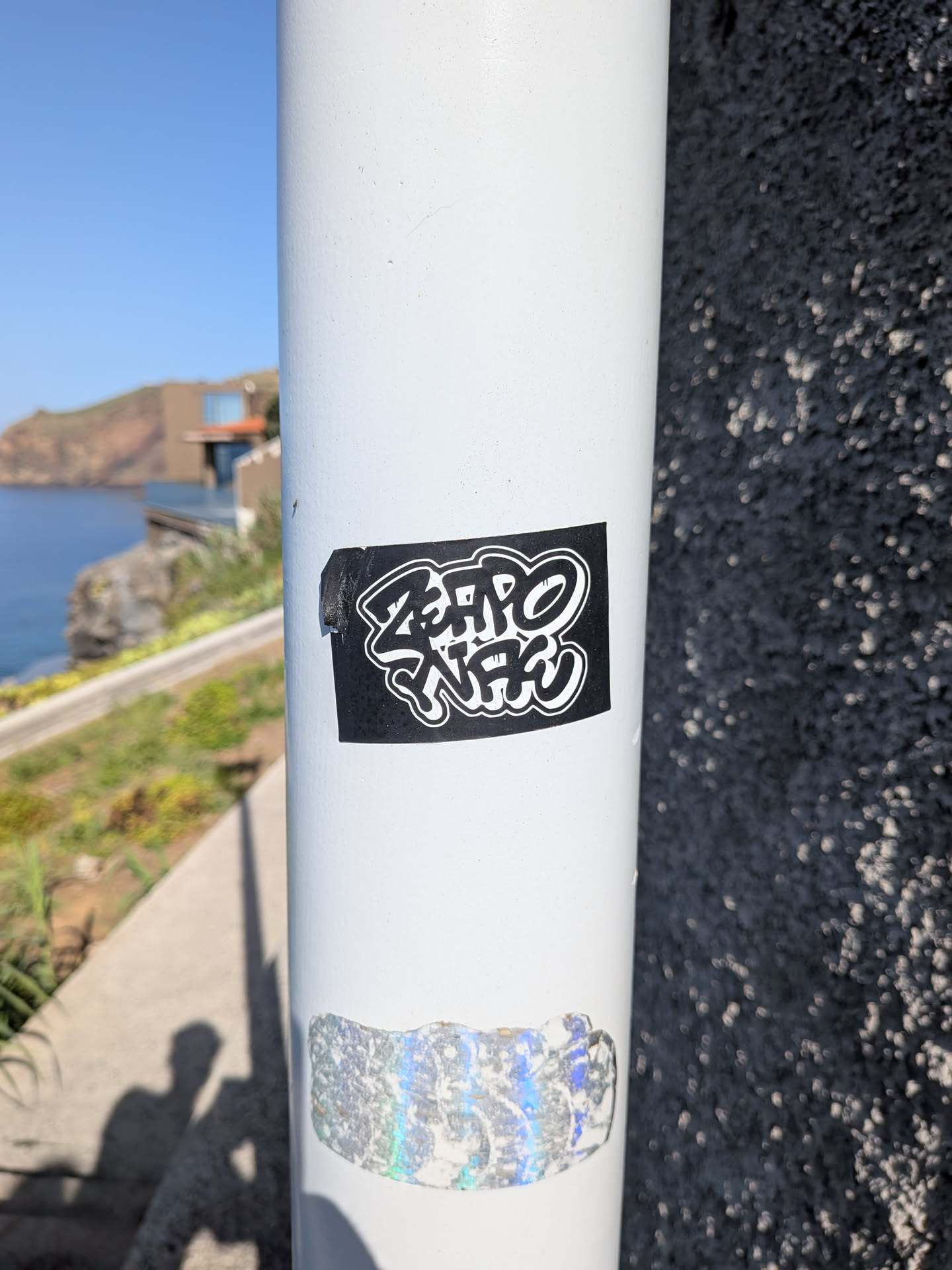A rectangular black sticker with stylized white graffiti lettering reading 'ZERO WAC' outlined in white, affixed to a white metal pole.
