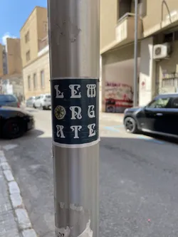Street sticker A rectangular sticker with a dark, textured background, appearing as a deep teal or distressed dark blue-green. It features highly stylized white capital letters arranged in three rows. The first row reads 'LEM'. The second row begins with a detailed illustration of a yellow and green lemon slice, followed by the letters 'N G'. The third row reads 'A T E'. The letters have an ornate, almost art nouveau or gothic-inspired design, with prominent serifs and decorative flourishes. The sticker shows visible signs of wear, including slight peeling at the edges and minor surface scuffs, indicating it is a physical sticker applied to the metal pole.