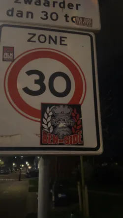 Street sticker The sticker is a square design with a black border. Its central figure is a stylized, angry-looking character with a head resembling a chess rook or a piston, colored in shades of grey. The character has prominent, clenched fists. The background is divided vertically: the left side features white or light grey wheat or laurel leaves, while the right side displays similar leaves in red. Below the character, the text 'BEN-SIDE' is prominently displayed in red letters with a white outline. A smaller 'AZ' is visible on the character's head.