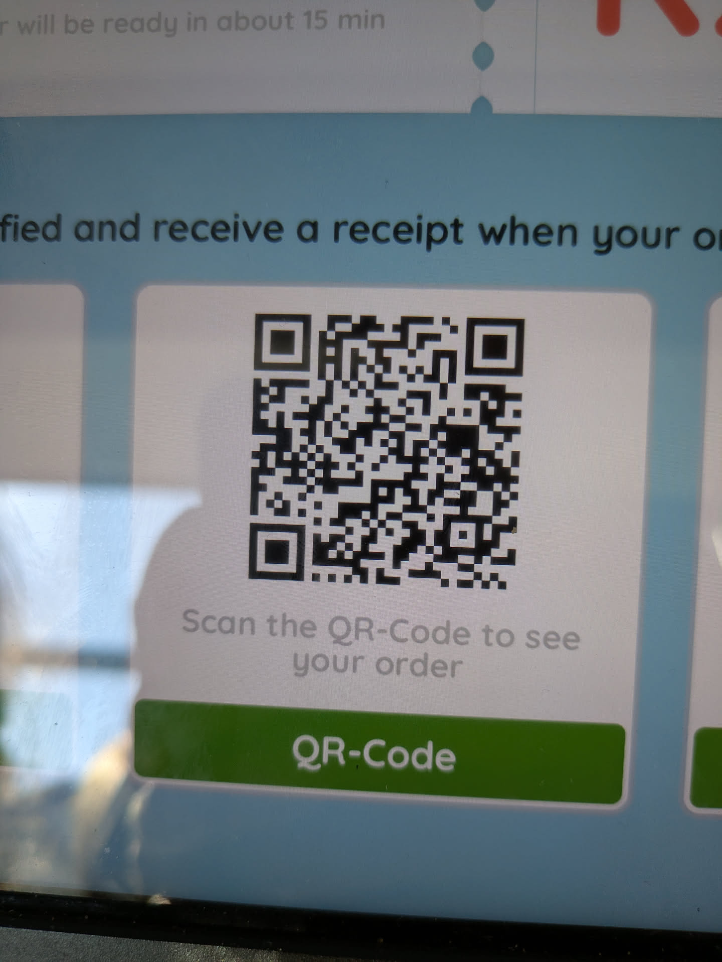 The image shows a close-up of a digital display screen, likely at a restaurant or food service establishment. The main focus is a large, square black and white QR code prominently centered on a light gray background. Below the QR code, there is text that reads "Scan the QR-Code to see your order." At the bottom of the screen, there's a green rectangular button with rounded corners, also labeled "QR-Code" in white text. Partial text is visible above the QR code, suggesting the context of order notification and receipt, such as "...fied and receive a receipt when your or...". Further up, some text indicates an estimated waiting time: "... will be ready in about 15 min". The screen has a clean, modern interface, and there are reflections on its surface from the surrounding environment.