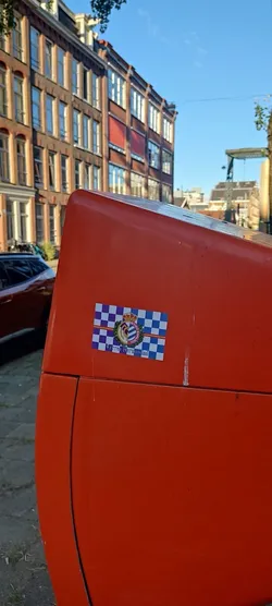 Street sticker The sticker is rectangular, featuring a prominent crest at its center. The background is a checkered pattern of purple and white squares, with a horizontal red stripe running through the middle. The crest itself is topped with a golden crown and features a shield with vertical blue and white stripes, possibly representing a sports club. It is encircled by a green and gold wreath. Text is visible above, below, and at the very bottom of the crest. The overall appearance is colorful and emblematic.