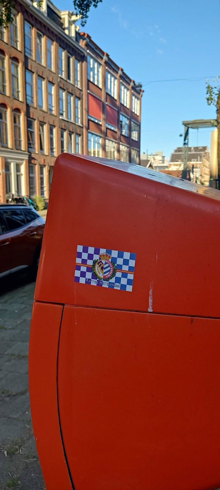 The sticker is rectangular, featuring a prominent crest at its center. The background is a checkered pattern of purple and white squares, with a horizontal red stripe running through the middle. The crest itself is topped with a golden crown and features a shield with vertical blue and white stripes, possibly representing a sports club. It is encircled by a green and gold wreath. Text is visible above, below, and at the very bottom of the crest. The overall appearance is colorful and emblematic.