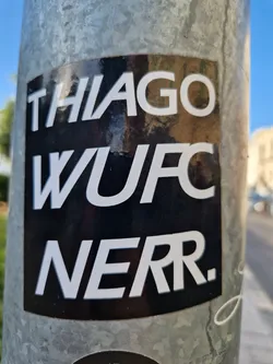 Street sticker A black and white sticker with the text "Thiago WUFC NERR." on a metal pole. The sticker has a simple design with bold, uppercase lettering.