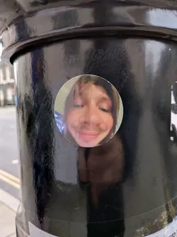 Street sticker A circular sticker featuring a close-up portrait of a person's face, heavily distorted by what appears to be a wide-angle or 'fish-eye' lens effect. The person has light brown hair, visible around the edges of the face, and is shown with closed eyes and a wide, exaggerated smile. The nose is flattened and widened due to the distortion, and the cheeks are prominent. The skin tone is fair, with hints of pink. The background within the sticker is muted, possibly an indoor setting, with some faint blue and purple reflections on the left side. A slight vertical crease is visible down the center of the sticker.