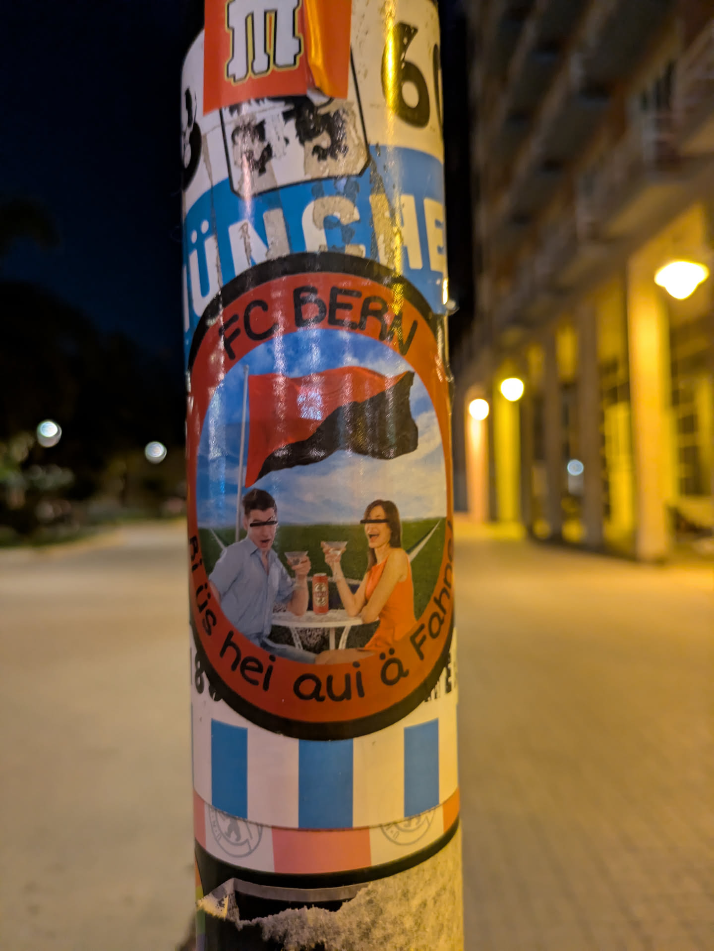 The sticker shows a man and woman at a table, toasting with drinks. In the background, there is a flag. The sticker has the words 'FC BERN' at the top and additional text around the bottom edge. It is stuck on a light pole.