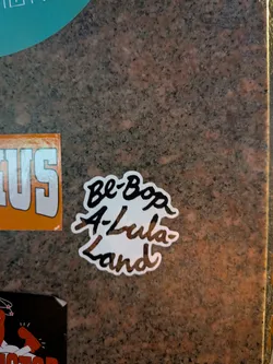 Street sticker The sticker is white with a black outline and features handwritten text. The text is arranged in three lines: 'Be-Bop' on the top, 'A-Lula' in the middle, and 'Land' on the bottom. The lettering is stylized and slightly playful, with a whimsical feel. The overall shape of the sticker is irregular, following the contours of the text.