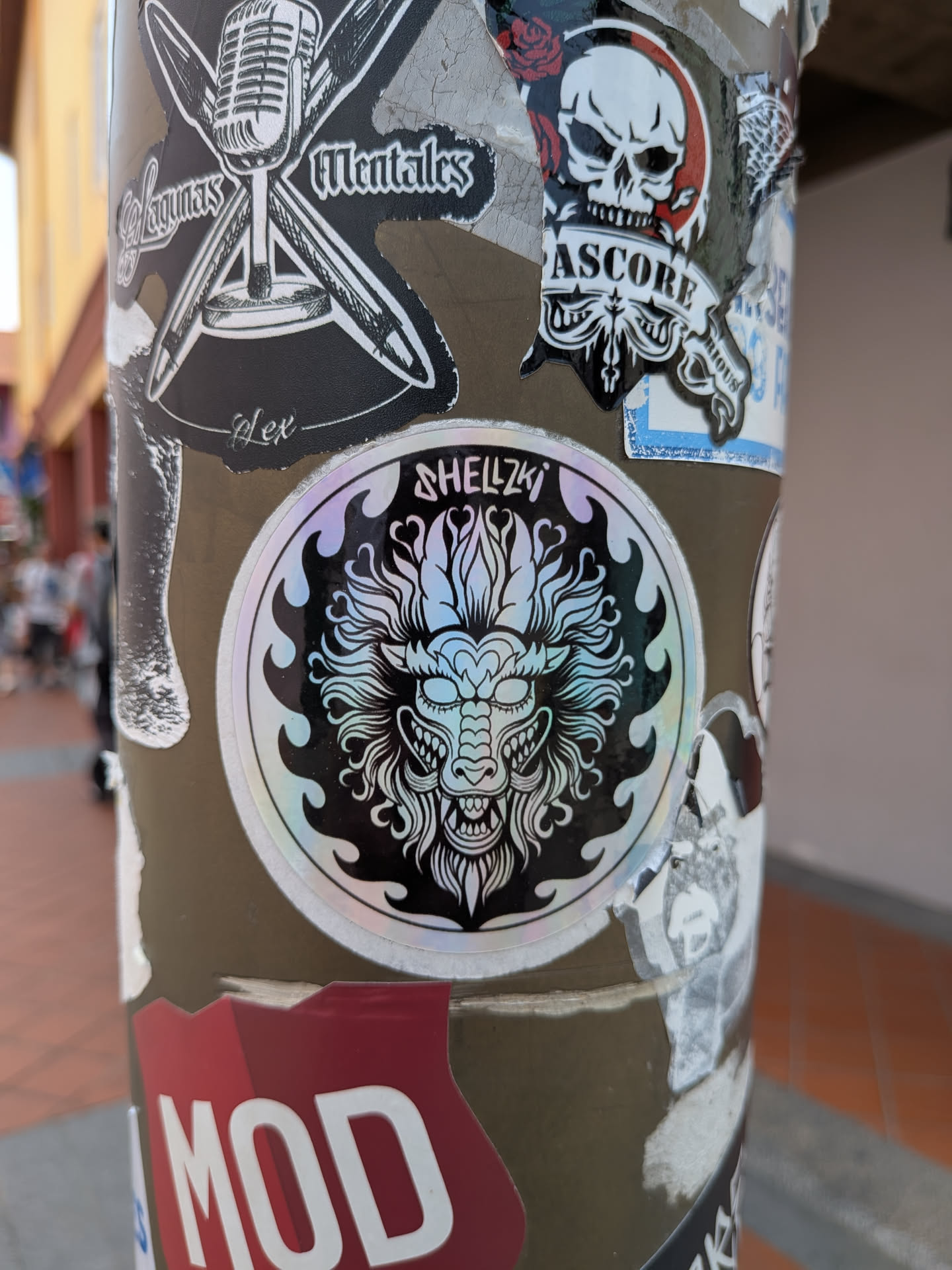 A circular sticker with a holographic, iridescent finish. It depicts a stylized, detailed dragon or guardian lion head in white outline against a black background, framed by flame motifs. The text 'SHELLZKI' is written at the top.