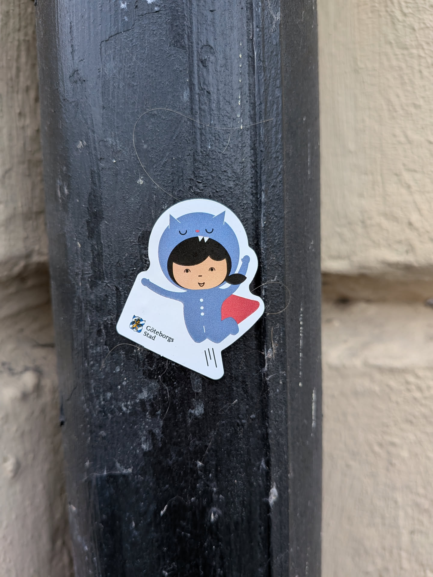 Street sticker The sticker features a cartoon illustration of a child wearing a blue hooded onesie designed to look like a cat, complete with ears and a face on the hood. The child has dark hair, brown eyes, and a cheerful expression. A small red cape is attached to the back of the onesie. In the lower left corner of the sticker, there is a small emblem with blue and yellow elements, and the text 'Göteborgs Stad' below it. The sticker has a white border and is die-cut to the shape of the illustration. There are also three thin vertical lines below the emblem.