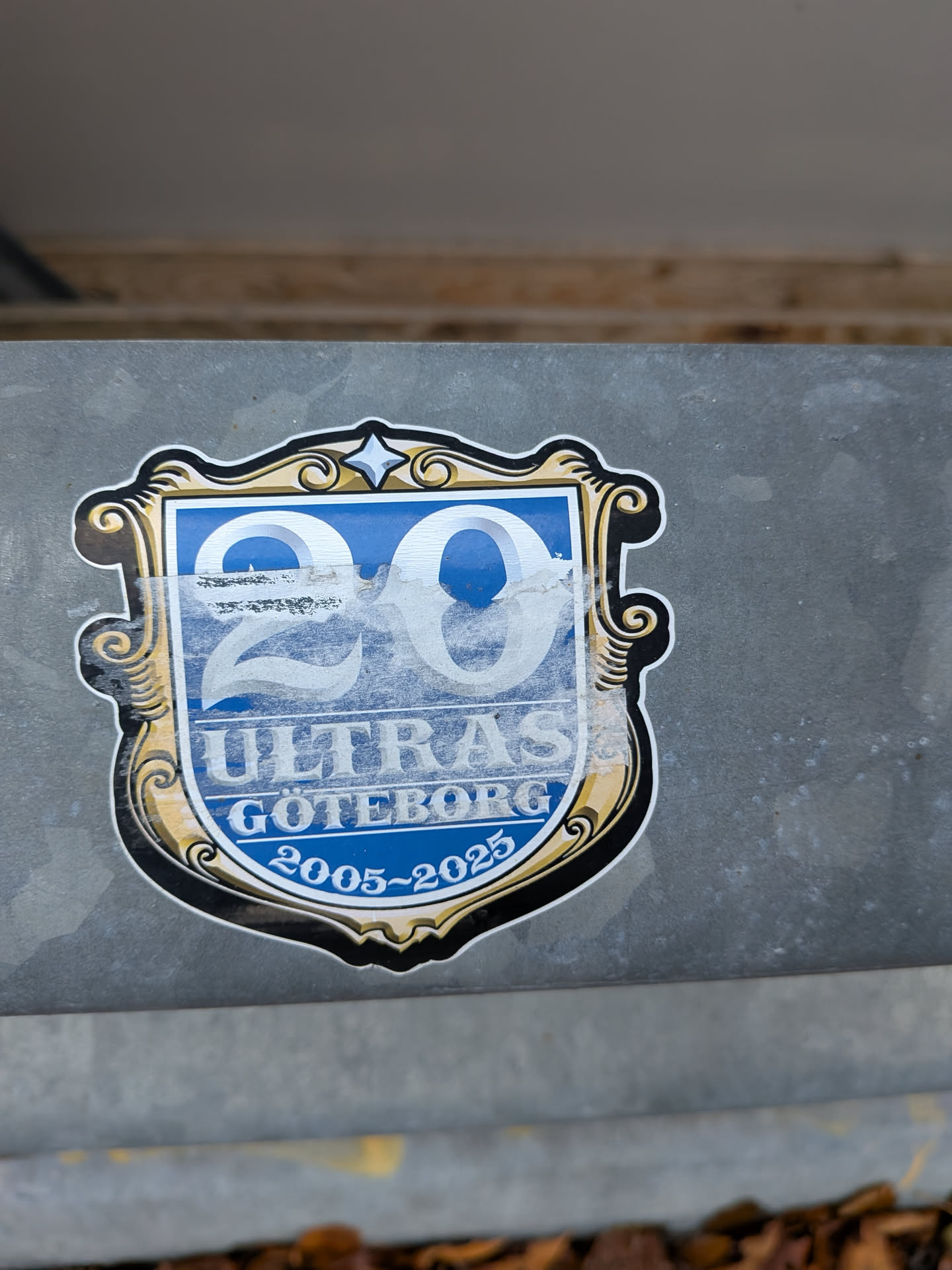 Street sticker The sticker is shield-shaped with a decorative, ornate border in black, gold, and white. The central area features a bright blue background. Prominently displayed are the large numbers '20' in a light, almost white color, with a subtle sheen. Below the '20', the word 'ULTRAS' is written in white capital letters. Beneath 'ULTRAS', 'GOTEBORG' is also in white capital letters. At the bottom of the shield, the text '2005~2025' is inscribed in white. The sticker shows some signs of wear, with a translucent, slightly scuffed layer over the '20' and parts of the blue background.