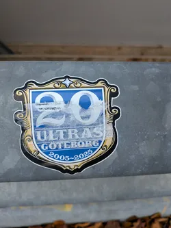 Street sticker The sticker is shield-shaped with a decorative, ornate border in black, gold, and white. The central area features a bright blue background. Prominently displayed are the large numbers '20' in a light, almost white color, with a subtle sheen. Below the '20', the word 'ULTRAS' is written in white capital letters. Beneath 'ULTRAS', 'GOTEBORG' is also in white capital letters. At the bottom of the shield, the text '2005~2025' is inscribed in white. The sticker shows some signs of wear, with a translucent, slightly scuffed layer over the '20' and parts of the blue background.