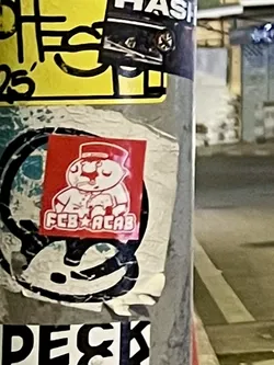 Street sticker A square-shaped sticker with a bright red background featuring a white, cartoonish character. The character appears to be a bear or similar animal, standing upright and wearing a red hat with a white band and a white uniform jacket with buttons. Its hands are clasped in front, and it has a small red nose. Below the character, in white capital letters, is the text 'FCB' followed by a star symbol, and then 'ACAB'. The sticker shows signs of wear, with slight creases and peeling edges, indicating it's been affixed for some time.