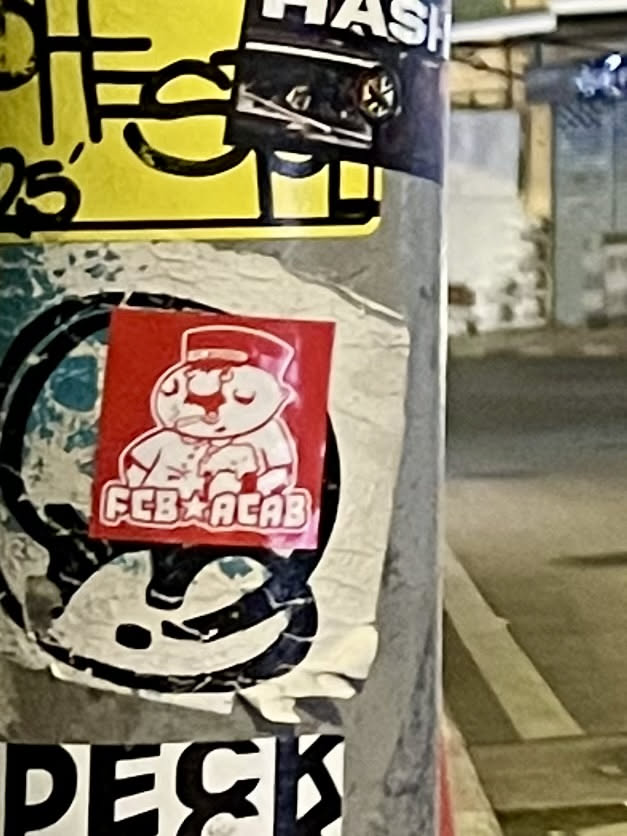 A square-shaped sticker with a bright red background featuring a white, cartoonish character. The character appears to be a bear or similar animal, standing upright and wearing a red hat with a white band and a white uniform jacket with buttons. Its hands are clasped in front, and it has a small red nose. Below the character, in white capital letters, is the text 'FCB' followed by a star symbol, and then 'ACAB'. The sticker shows signs of wear, with slight creases and peeling edges, indicating it's been affixed for some time.