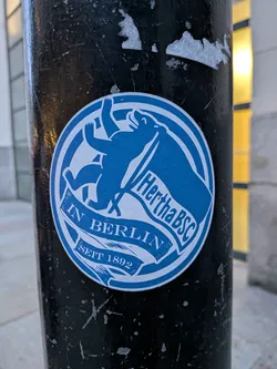 Street sticker A circular sticker featuring a stylized blue bear, the iconic symbol of Berlin, standing on its hind legs with an open mouth, holding a flowing banner. The banner prominently displays 'Hertha BSC' in white text. Below the bear, another banner segment reads 'IN BERLIN', and at the very bottom, a smaller segment states 'SEIT 1892'. The entire design is framed by multiple concentric blue and white rings, giving it the appearance of a club crest or emblem. The sticker is predominantly blue and white, with the blue elements contrasting against the white background.