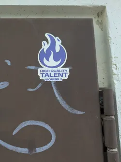 Street sticker The sticker is predominantly white with blue accents. It features a stylized flame design in blue and white. Below the flame, the text "HIGH QUALITY TALENT" is printed in a simple font. Below that, the word "WORKSIBLE" is also printed. The sticker is adhered to a brown metal surface that has some white painted markings.