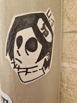 Street sticker A black and white sticker featuring a stylized skull character with stitches on its face and a unique headdress. The sticker seems to be hand-drawn and has a slightly grungy texture. The artist's initials (or a tag) 'U.S' are visible on the top right corner of the sticker.