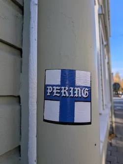 Street sticker The sticker is rectangular with rounded corners and a black border. It features a design resembling the Finnish flag, with a blue Nordic cross on a white background. The word 'PEKING' is prominently displayed in the center of the cross, in a stylized white font with a black outline. The sticker appears to be slightly textured and adhered to a light-colored pole.