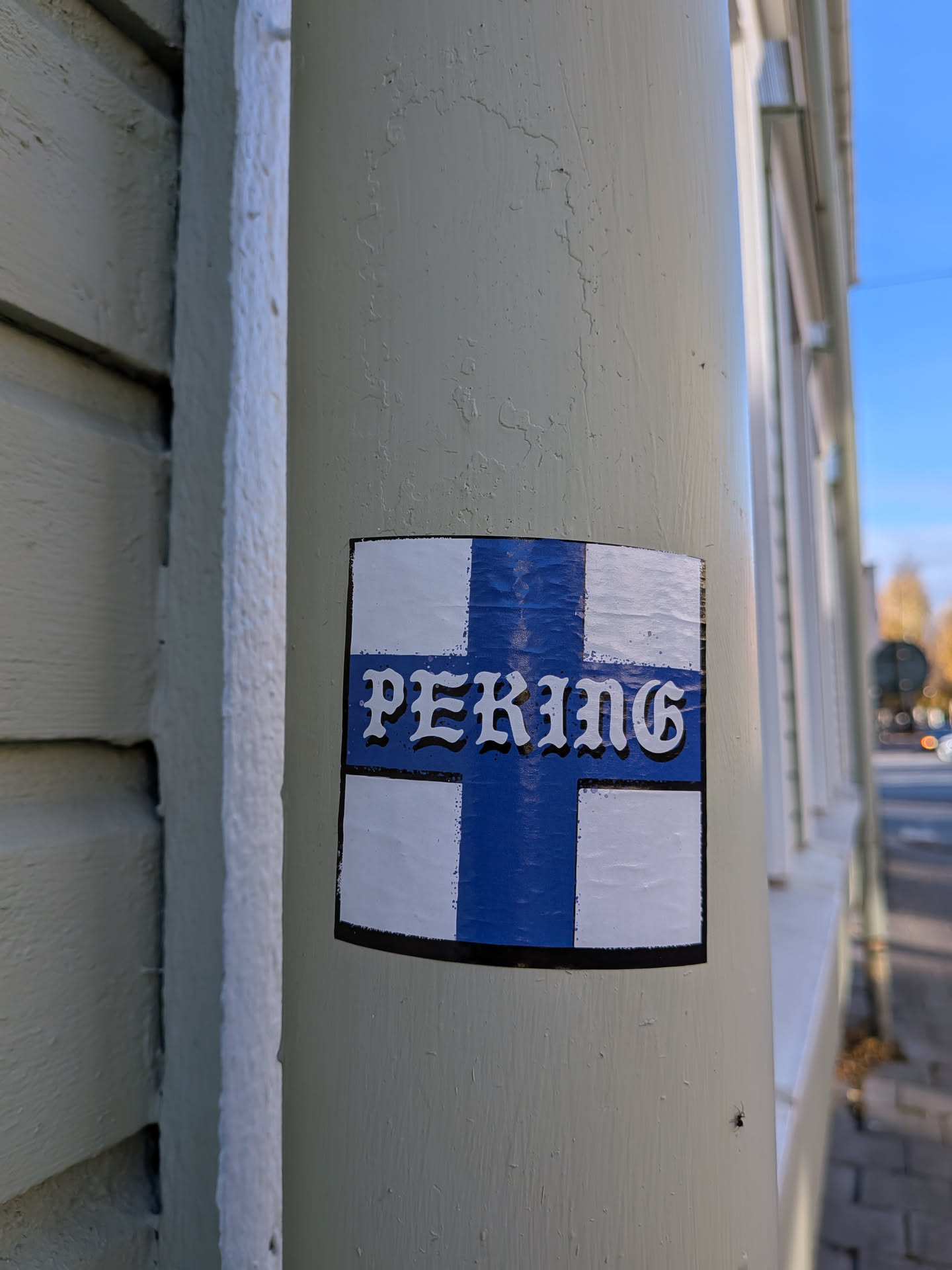 The sticker is rectangular with rounded corners and a black border. It features a design resembling the Finnish flag, with a blue Nordic cross on a white background. The word 'PEKING' is prominently displayed in the center of the cross, in a stylized white font with a black outline. The sticker appears to be slightly textured and adhered to a light-colored pole.