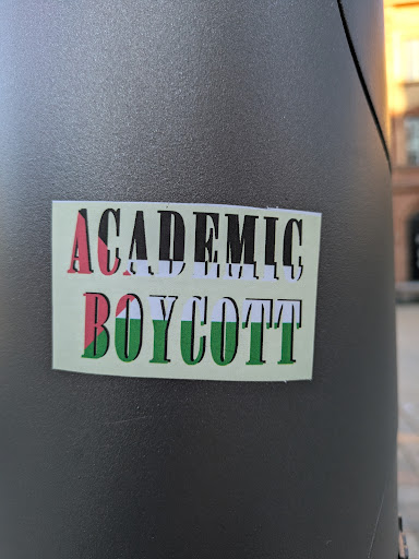 A rectangular sticker adhered to a gray metal post. The sticker reads "ACADEMIC BOYCOTT" in bold, block letters. The letters are filled with a pattern of red, black, white, and green, colors often associated with Palestinian solidarity. The background of the sticker is light green.